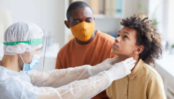 Portrait of male doctor examining African-American boy and palpating throat during consultation or covid in hospital